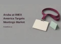 Conceptual editorial illustration of a blank conference badge on a burgundy lanyard beside a mint tent card, with the headline Aruba at IMEX America Targets Meetings Market and ArubaNews.ca branding.