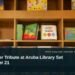 Illustrative stock photo for “Knipscheer Tribute at Aruba Library Set for October 21”: Children’s books arranged on library shelves. Photo by Zoshua Colah via Unsplash; not a photograph of the specific event. Headline and ArubaNews.ca branding appear in a lower-third news tile.