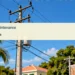 Conceptual editorial illustration; a generic residential electrical distribution pole with insulated wires against clear Caribbean sky, upward perspective, simple houses softly out of focus, crisp blue and cream palette. Not a documentary image of the reported event.
