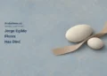 Conceptual editorial illustration of two ivory stones and a curved clay-coloured paper ribbon on a blue-grey surface, with the headline Jorge Egildo Flores Has Died and ArubaNews.ca branding.
